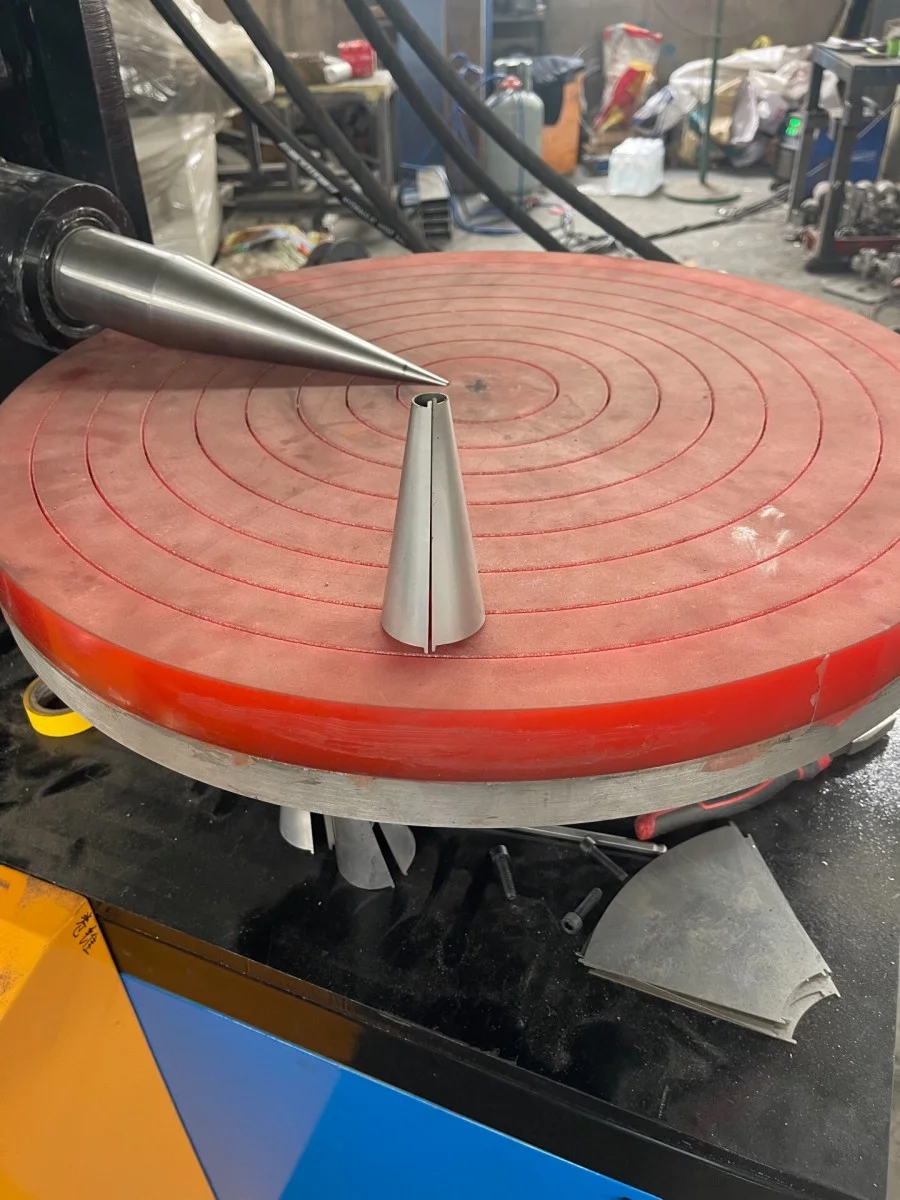 Крупный план рабочего стола конусопрокатного станка дискового типа, демонстрирующий заготовку веерообразной формы из нержавеющей стали и небольшую полуфабрикатную коническую трубу.